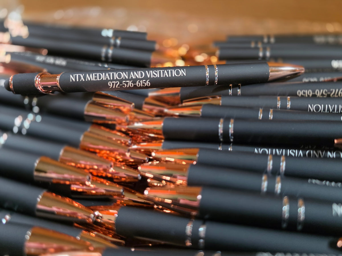 A close-up of multiple black pens with shiny rose-gold metal accents and laser engraved text arranged in a pile.