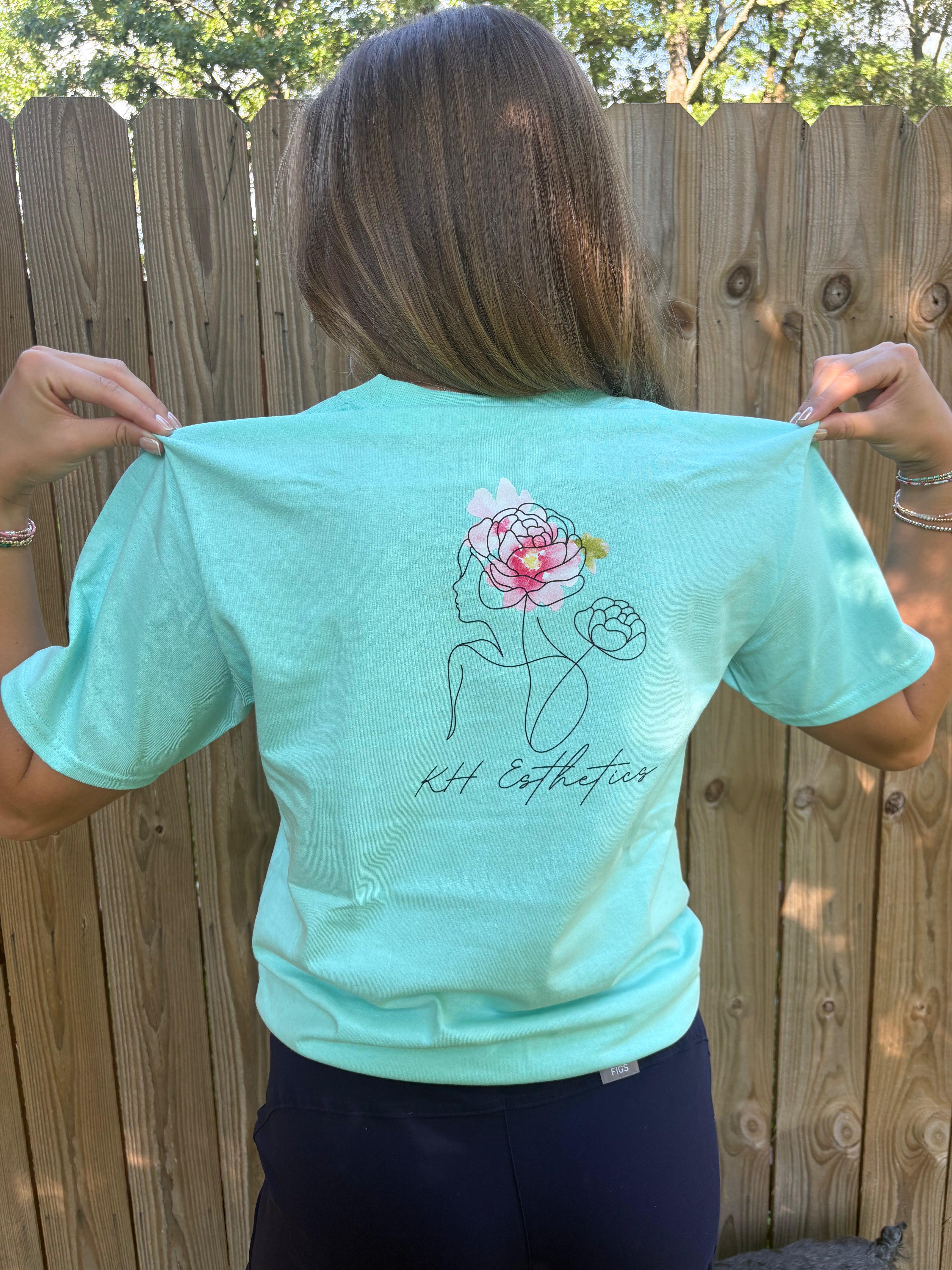 Person wearing a light blue t-shirt with a design and text on the back, standing in front of a wooden fence.